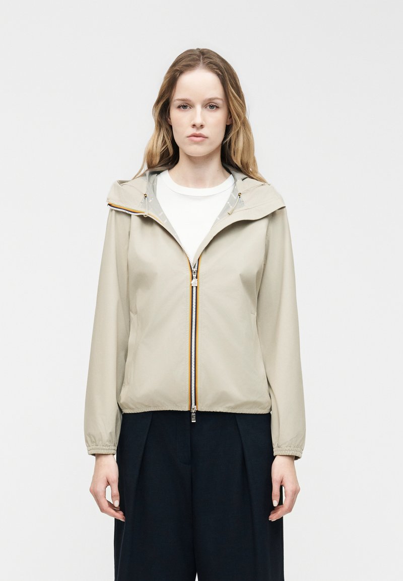 Νεαρή γυναίκα που φοράει μπεζ μπουφάν με φερμουάρ και κουκούλα πάνω από λευκό πουκάμισο και φαρδιά μαύρη παντελόνια, στέκεται μπροστά σε απλό ανοιχτό φόντο.