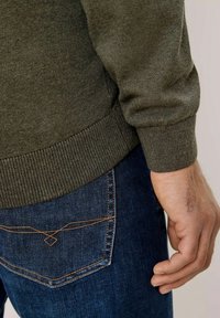 Pull en tricot vert foncé avec des poignets côtelés, accompagné d'un jean bleu foncé avec des surpiqûres contrastées sur la poche arrière.