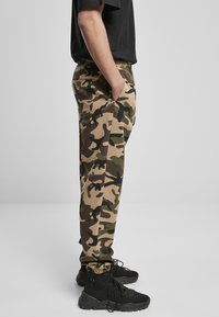 Beige, zöld és fekete mintázatú kamuflázs joggerek, elasztikus derékkal és zsebekkel, fekete sportcipővel párosítva.
