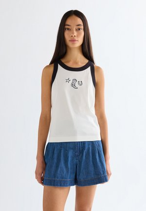 Νέα γυναίκα που φοράει λευκό αμάνικο τοπ με μαύρα περιθώρια και εικονίδια western θέματος, συνδυασμένο με μπλε τζιν σορτς, στέκεται μπροστά σε λευκό φόντο.