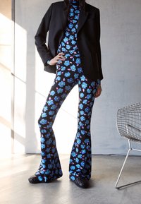 Pantalones acampanados y un suéter de cuello alto en tela negra con un patrón floral azul, combinados con una chaqueta negra. Unos zapatos negros texturizados completan el atuendo.