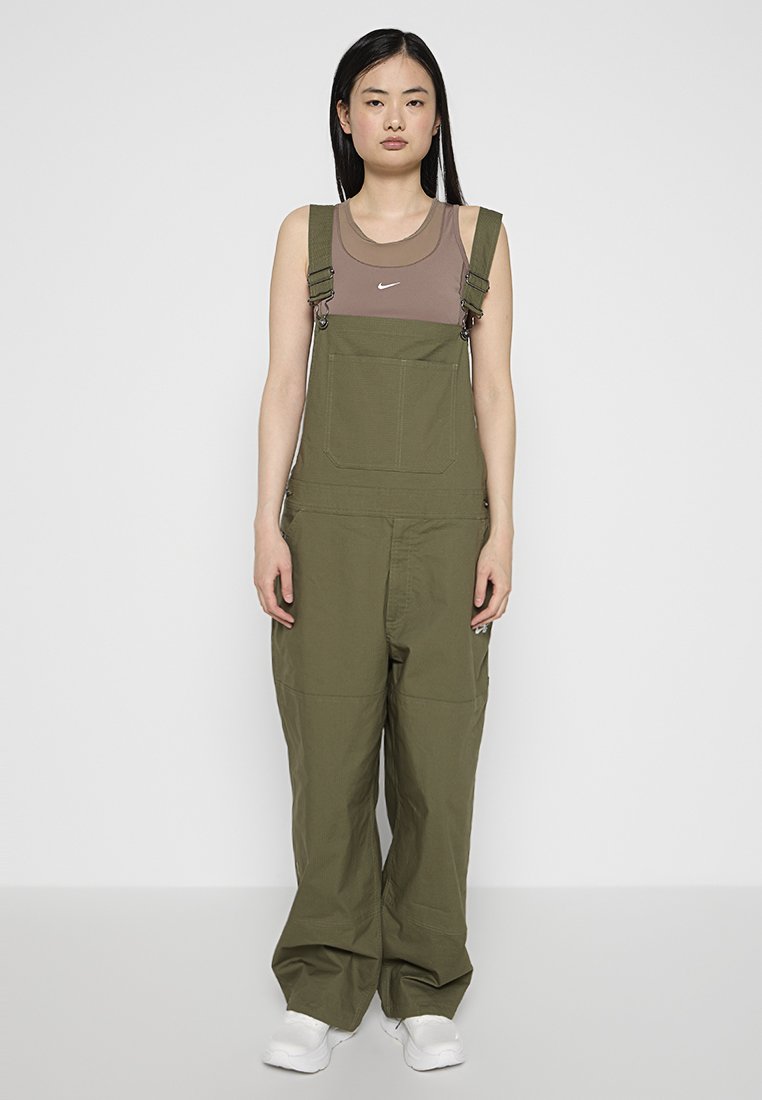 Olivegrüne Latzhose mit verstellbaren Trägern, großer Fronttasche und lässiger Passform. Kombiniert mit einem braunen Tanktop und weißen Turnschuhen.