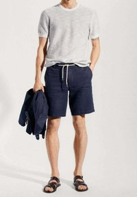 Chemise à manches courtes gris rayé, short marine avec cordon de serrage, tenant une veste marine, et portant des sandales noires. Tissu lisse et coupe décontractée.