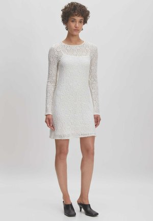 Weißes Spitzenkleid mit langen Ärmeln, strukturiertem Stoff, Rundhalsausschnitt und leicht ausgestellt Saum. Kombiniert mit schwarzen offenen Mules.