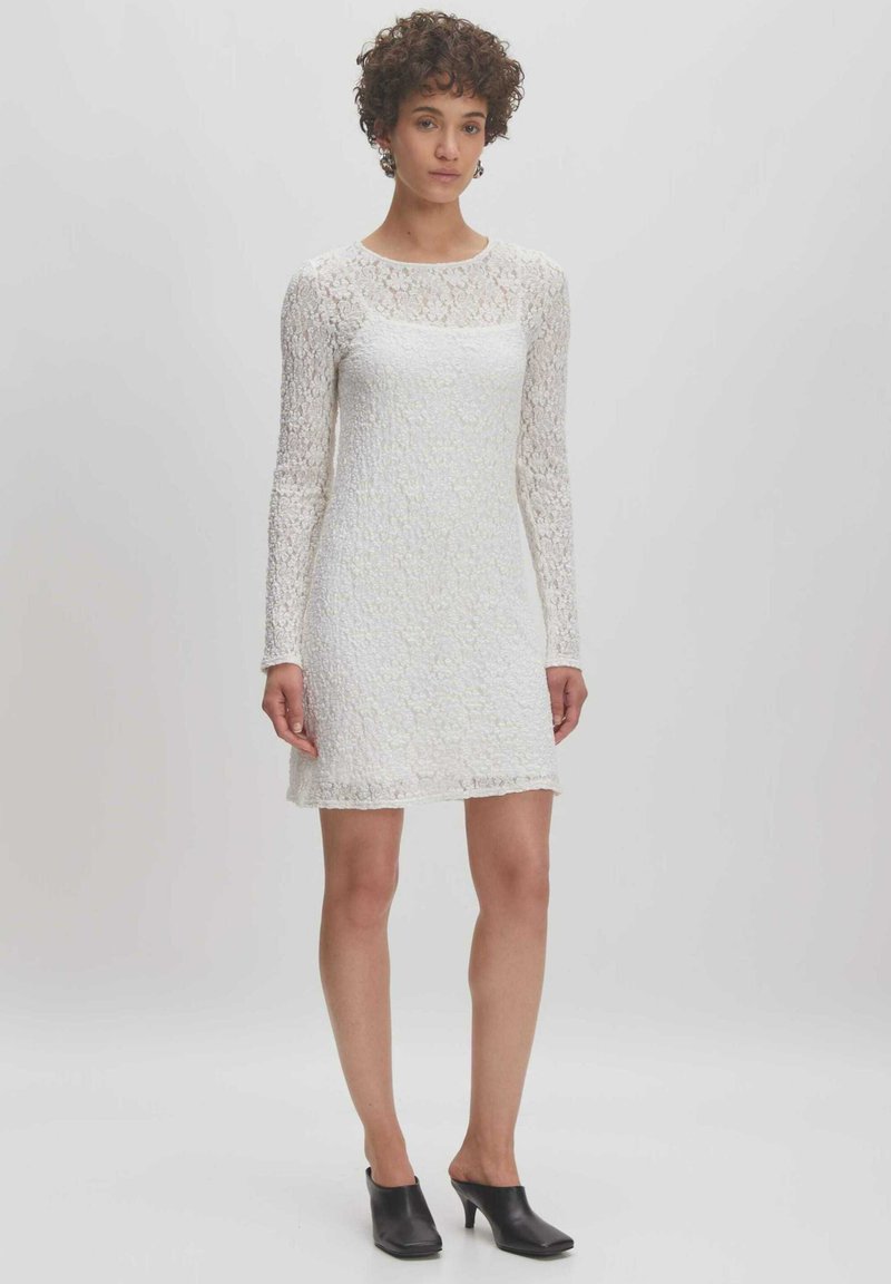 Weißes Spitzenkleid mit langen Ärmeln, strukturiertem Stoff, Rundhalsausschnitt und leicht ausgestellt Saum. Kombiniert mit schwarzen offenen Mules.