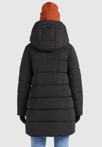 Persona con un abrigo de invierno acolchado negro con capucha, gorro de punto naranja, guantes negros y jeans azul claro, visto desde atrás.