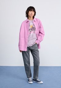 Oversized pink corduroy shirt with front buttons over a light purple graphic t-shirt. Worn with gray straight-leg jeans and navy sneakers.