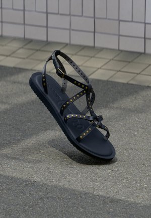 Sandalia azul marino con tiras grises tachonadas, suela plana y hebilla ajustable. Presenta una plantilla texturizada y un diseño minimalista.