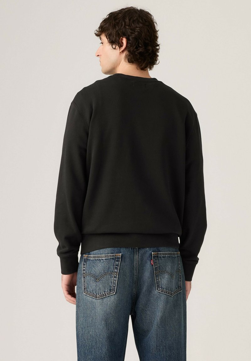 Homme aux cheveux bouclés portant une chemise noire à manches longues et un jean bleu, debout de dos contre un fond clair uni.