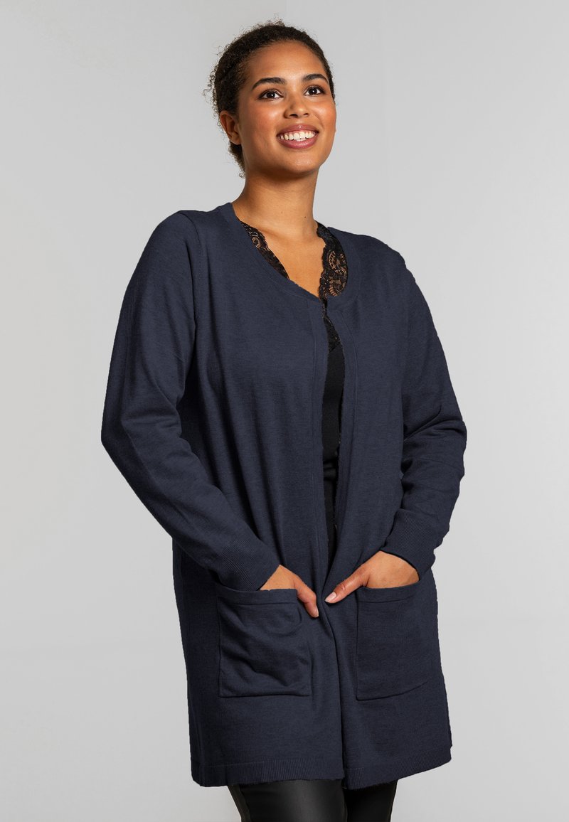 Hymyilevä nainen, jolla on pitkä laivaston sininen neuletakki taskuilla mustan pitsikoristeisen paidan päällä, seisomassa yksinkertaisen vaaleanharmaan taustan edessä.