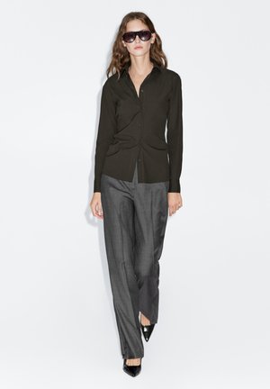 Olive green button-up shirt paired with tailored gray trousers and black pointed heels. Model also wears dark sunglasses. Smooth fabric texture.