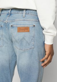 Jeans in denim azzurro con un'etichetta in pelle marrone Wrangler, tasche posteriori visibili e dettagli di cucitura a contrasto.