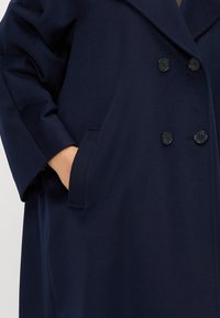 Cappotto doppiopetto blu navy realizzato in tessuto testurizzato, dotato di bottoni grandi, colletto a scialle e tasche laterali.