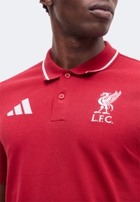 Homme portant un polo rouge avec un logo Adidas blanc et l'emblème de l'oiseau du Liverpool FC sur la poitrine, col boutonné.
