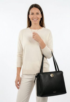 Femme portant un pull crème et un pantalon, souriante, tenant un sac à main en cuir noir avec double anses et une sangle à détail doré sur le bras.