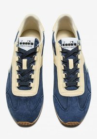 Sneakers di pelle scamosciata blu navy con dettagli crema, lacci testurizzati e suola in gomma. Presentano un logo prominente sulla linguetta.