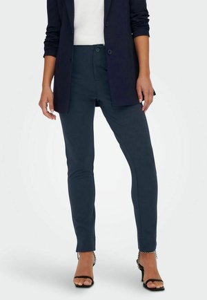 Mujer con pantalones ajustados azul oscuro, blazer negro, top blanco y sandalias negras de tacón con correas de cadena en el tobillo, posando frente a un fondo liso.