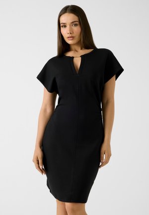 Femme aux cheveux longs et lisses portant une robe noire longueur genoux avec des manches courtes et une petite ouverture à l'avant du col avec un fermoir doré.