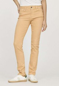 Jean slim beige clair avec cinq poches, une fermeture à bouton et à zip, et des coutures décoratives, associé à des baskets blanches.