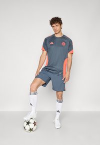 Grå idrettsjakke med neongule detaljer, kombinert med matchende shorts, hvite sokker med svarte striper, og fotballsko på en fotball.