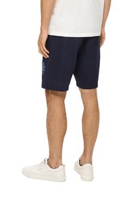 Shorts texturés bleu marine avec un motif floral sur la cuisse gauche, associés à des baskets blanches. Jambes visibles et t-shirt blanc.