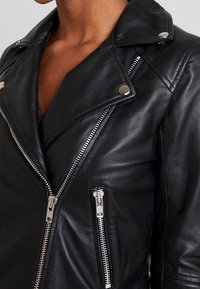 Veste en cuir noir de style biker avec des fermetures éclairs argentées, des boutons-pression et un design asymétrique à l'avant. Texture lisse et silhouette ajustée.