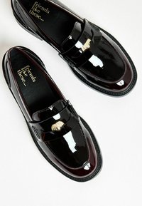 Mocasines de charol negro con un acabado brillante, que cuentan con un acento de león dorado en la correa y una suela burdeos en contraste.