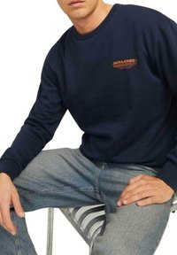 Sudadera azul marino con cuello redondo y mangas largas, que presenta un logo naranja en el lado izquierdo del pecho, combinada con jeans de tono azul claro.