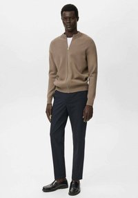 Man in een beige zip-up cardigan, wit onderhemd, zwarte op maat gemaakte broek en zwarte instappers, staand tegen een effen achtergrond.