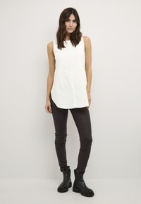 Top bianco senza maniche con abbottonatura, abbinato a pantaloni aderenti marrone scuro e stivaletti neri. Tessuto liscio e vestibilità comoda.