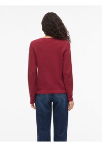 Γυναίκα με σγουρά μαλλιά που φοράει ένα υφασμάτινο μπορντό πουλόβερ και σκούρα μπλε τζιν, με θέα από πίσω σε λευκό φόντο.