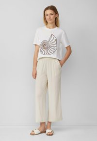 Witte t-shirt met een graphic van een spiraalschelp, gecombineerd met crèmekleurige wijde broek en witte sandalen met een gouden accent.