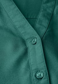 Camicia button-up color teal in tessuto dalla texture liscia, con scollatura a V e bottoni a contrasto con un logo impresso su di essi.