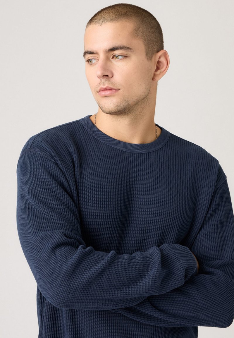 Marineblauer Langarmpullover aus Strick mit strukturiertem, Waffelmuster; runder Halsausschnitt und entspannte Passform; sichtbare Nähte an Schultern und Bündchen.