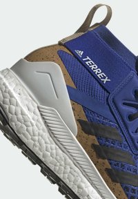 Gros plan d'une chaussure de trail running Adidas Terrex bleue et marron avec des bandes noires, une semelle blanche texturée et une languette beige au talon.