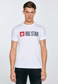 Jonge man met kort bruin haar die een wit "BIG STAR" T-shirt met een rood sterlogo draagt, staand tegen een effen lichte achtergrond.
