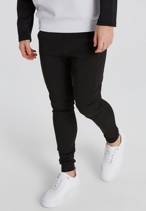 Schwarze sportliche Jogginghosen mit einer taillierten Passform, aus strukturiertem Material und mit einem gestickten Logo am Oberschenkel, kombiniert mit weißen Sneakers.