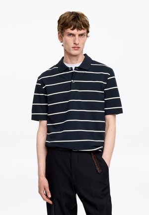 Jeune homme portant un polo noir avec des rayures horizontales blanches, un pantalon noir et une ceinture tressée, debout devant un fond blanc uni.