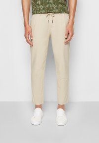 Pantaloni beige a sigaretta con vita regolabile a cordino e tasche laterali, abbinati a una camicia verde a maniche corte con stampa floreale e sneakers bianche.