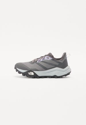 Zapatilla deportiva gris con parte superior de malla transpirable, suela de goma negra y acentos morados. Cuenta con una entresuela acolchada y un sistema de cordones.