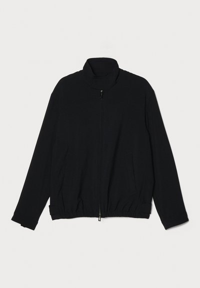 Casaco preto com fecho éclair, apresenta um colarinho alto, cintura elástica, tecido texturizado e dois bolsos laterais. Acabamento suave com um corte descontraído.