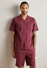 Burgundi spalvos trumpas rankovinis marškinėliai su tekstūruotu raštu, mygtukais priekyje ir dviem krūtinės kišenėmis; derinama su atitinkamais šortais iš tos pačios medžiagos.