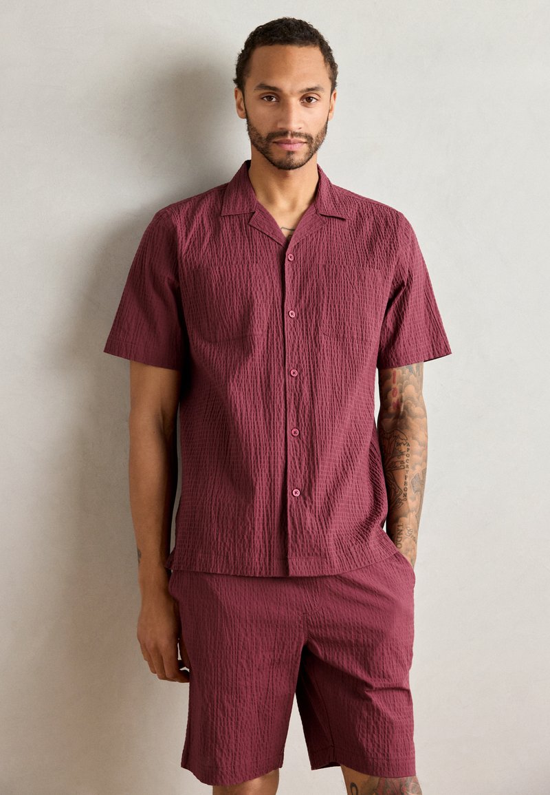 Burgundi spalvos trumpas rankovinis marškinėliai su tekstūruotu raštu, mygtukais priekyje ir dviem krūtinės kišenėmis; derinama su atitinkamais šortais iš tos pačios medžiagos.