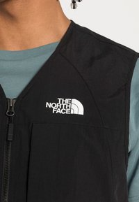 Czarna bezrękawnik z zamek błyskawicznym i białym logo The North Face. Noszony na niebieskiej koszuli z krótkim rękawem, charakteryzującej się gładką fakturą.