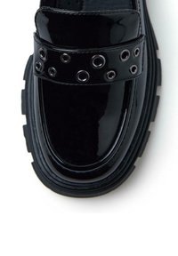 Mocasines de charol negro con una correa ancha que presenta ojales metálicos, suela de goma elevada y un acabado suave y brillante.