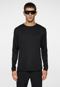 Svart långärmad skjorta i slätt tyg, med rundad halsringning och minimalistisk design. Bärs med eleganta svarta solglasögon.