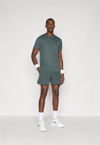 Conjunto atlético incluindo uma camisa de manga curta verde escuro e calções combinando. Os acessórios incluem braceletes brancos e sapatilhas desportivas.