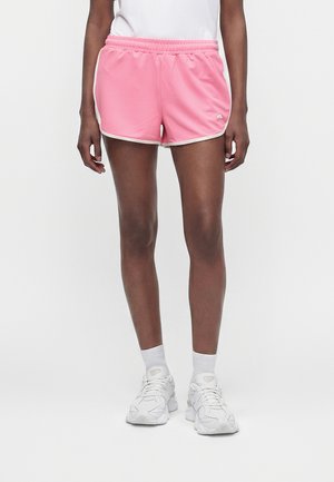 Person iført lyserøde træningsshorts med hvide kanter, hvide sokker og hvide sneakers, stående mod en ensfarvet baggrund.