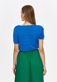 Niebieska dziergana koszula z krótkim rękawem z ażurowym wzorem; połączona z luźną, zieloną spódnicą plisowaną. Widok z tyłu ukazuje zaokrąglony dekolt.