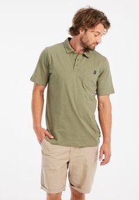 Olijfgroen polo-shirt met een klassieke kraag, een drie-knopen sluiting, een zak op de linkerborst en een zachte textuur. Draagt met beige shorts.